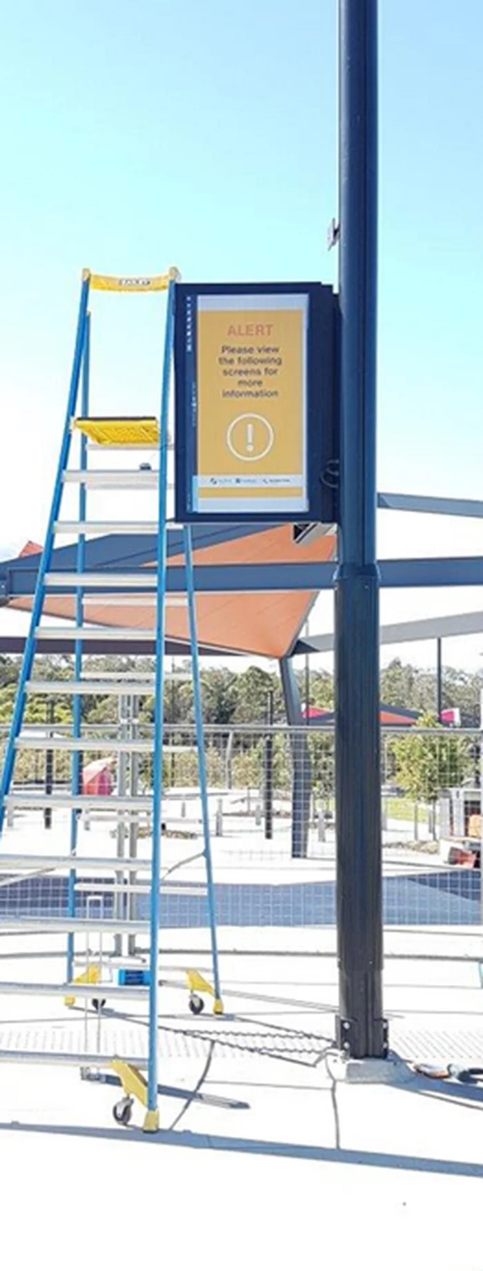 Pole mounted weather rated display carrying a safety message at an open air recreation centre, with an access platform beside it
